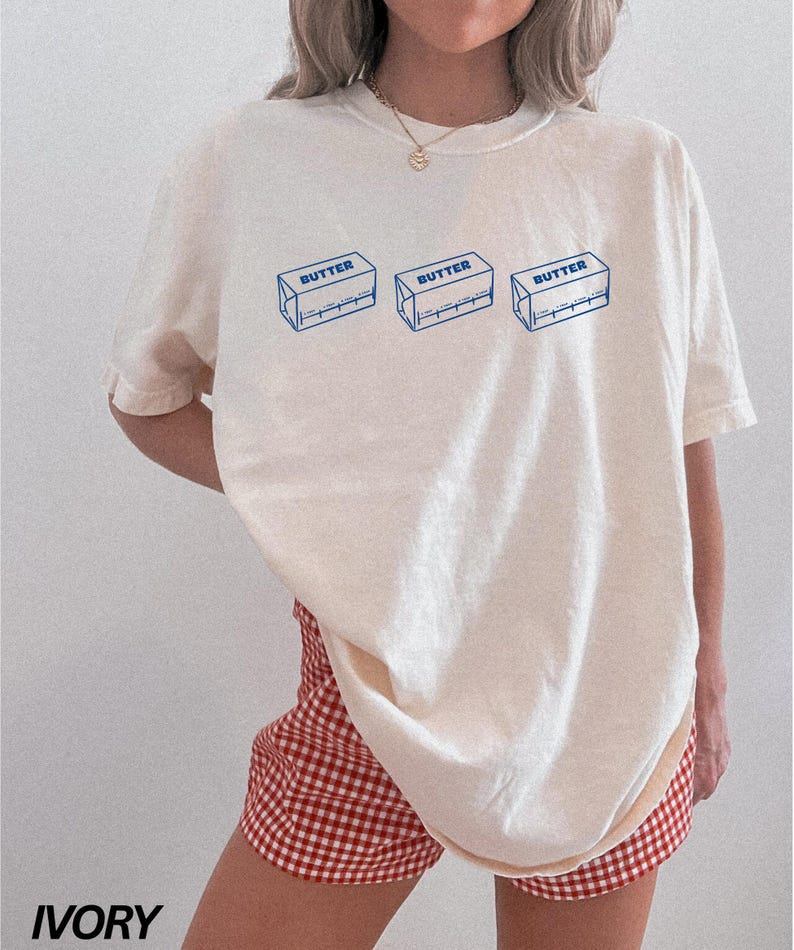 Puede incluir: Una camiseta blanca de gran tama&ntilde;o con un gr&aacute;fico de tres envases de mantequilla en barra con la palabra "BUTTER" impresa en cada uno. La camiseta la lleva puesta una persona que lleva pantalones cortos de cuadros rojos y blancos.