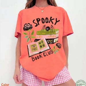 Op de afbeelding: Felzalmroze T-shirt met een griezelig boekenclubontwerp. De afbeelding bevat een spin op boeken, rozen en de woorden "Spooky Book Club". Het shirt wordt gecombineerd met een roze en witte geruite short.