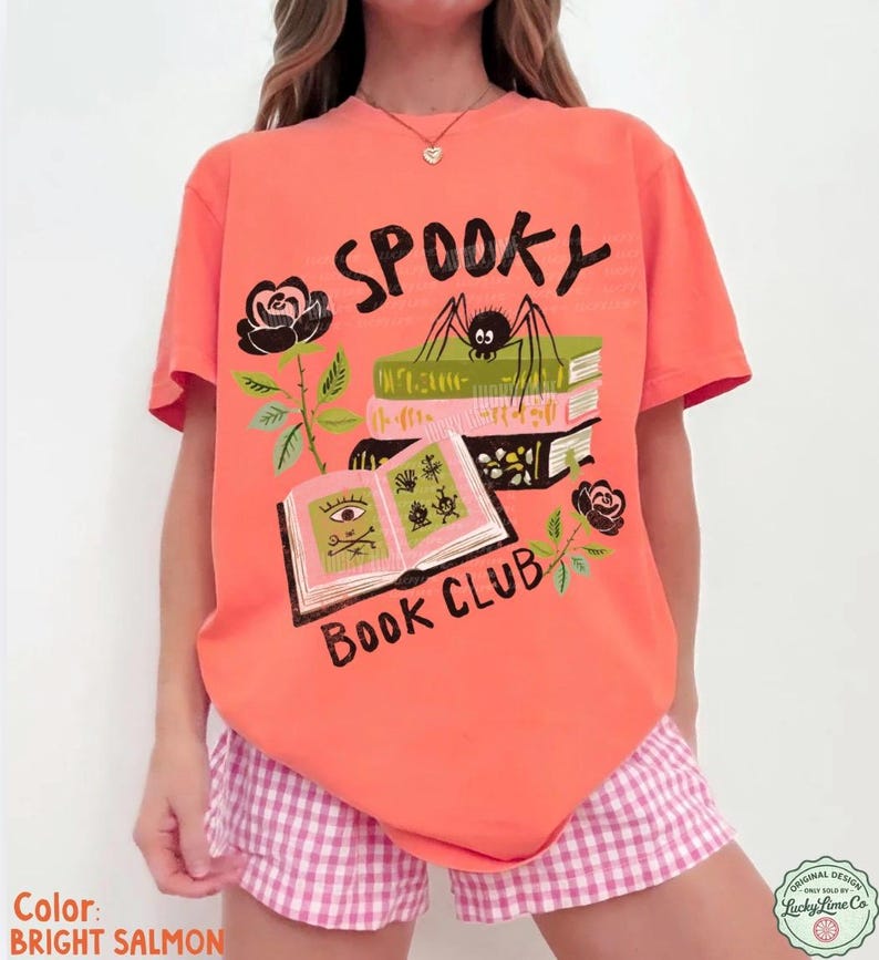 Op de afbeelding: Felzalmroze T-shirt met een griezelig boekenclubontwerp. De afbeelding bevat een spin op boeken, rozen en de woorden "Spooky Book Club". Het shirt wordt gecombineerd met een roze en witte geruite short.