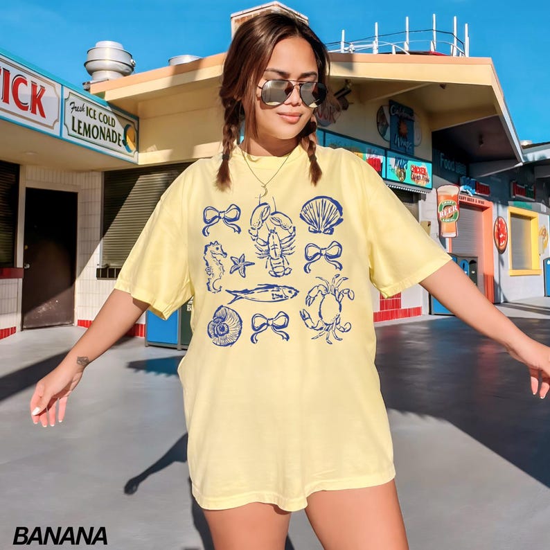 May include: A yellow t-shirt with a blue graphic print of various sea creatures and bows. The graphic includes a seahorse, a starfish, a lobster, a seashell, a crab, and a bow.