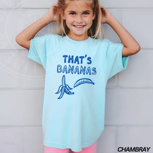 Op de afbeelding: Een lichtblauw T-shirt met de tekst "THAT'S BANANAS" in blauwe letters. Het shirt heeft een blauwe illustratie van twee bananen, een gepeld en een heel.
