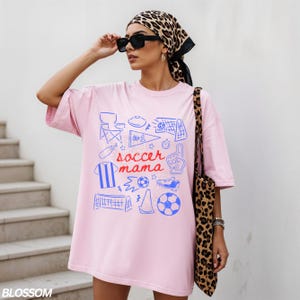 May include: A light pink t-shirt with a blue soccer-themed graphic and the words "soccer mama". The graphic includes a soccer ball, goal, and other soccer-related icons. The person is wearing a leopard print scarf and sunglasses.
