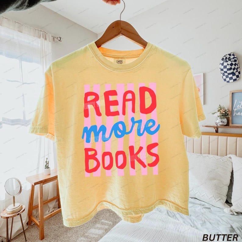 K&ouml;nnte beinhalten: Ein blassgelbes Kurzarm-T-Shirt mit einem Grafikdesign aus vertikalen rosa Streifen und dem Text "READ more BOOKS" in roter und blauer Schrift. Das T-Shirt h&auml;ngt auf einem Holzkleiderb&uuml;gel vor einem hellen Schlafzimmerhintergrund.