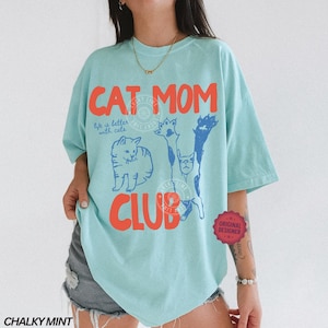 Puede incluir: Una camiseta azul claro con las palabras "CAT MOM CLUB" en naranja y azul. La camiseta presenta ilustraciones de gatos y la frase "life is better with cats". La camiseta tiene un corte holgado.