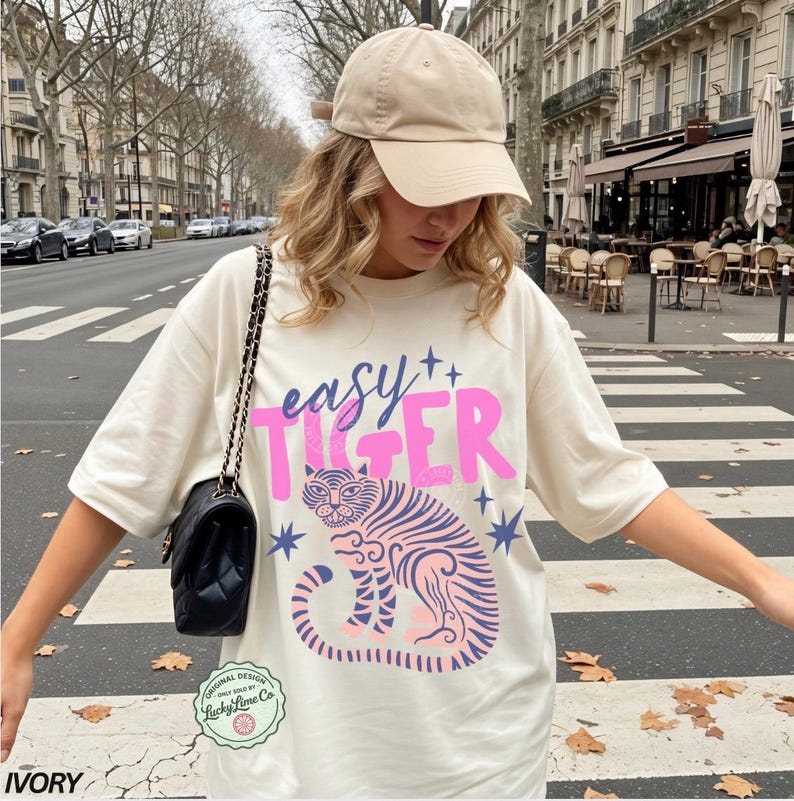 Puede incluir: Camiseta de color marfil con el texto "easy TIGER" en rosa y azul, con una ilustraci&oacute;n de tigre. La camiseta se combina con una gorra de b&eacute;isbol beige y un bolso de hombro negro acolchado. La imagen est&aacute; tomada al aire libre.