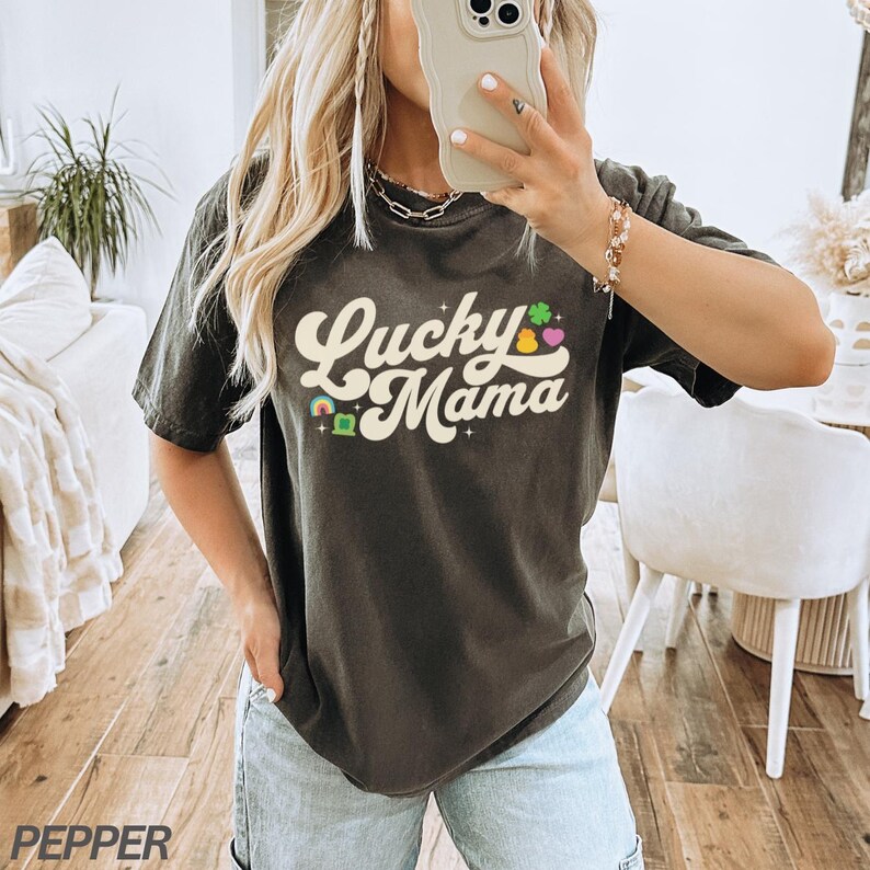 Peut inclure: Un t-shirt noir avec le texte "Lucky Mama" dans une police r&eacute;tro. Le texte est entour&eacute; de tr&egrave;fles, de c&oelig;urs et d'arcs-en-ciel color&eacute;s.