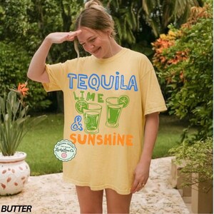 May include: A butter yellow t-shirt with the words "TEQUILA LIME & SUNSHINE" in blue, green, and orange. The shirt features two cocktail glasses with lime slices. The t-shirt is a casual, oversized fit.