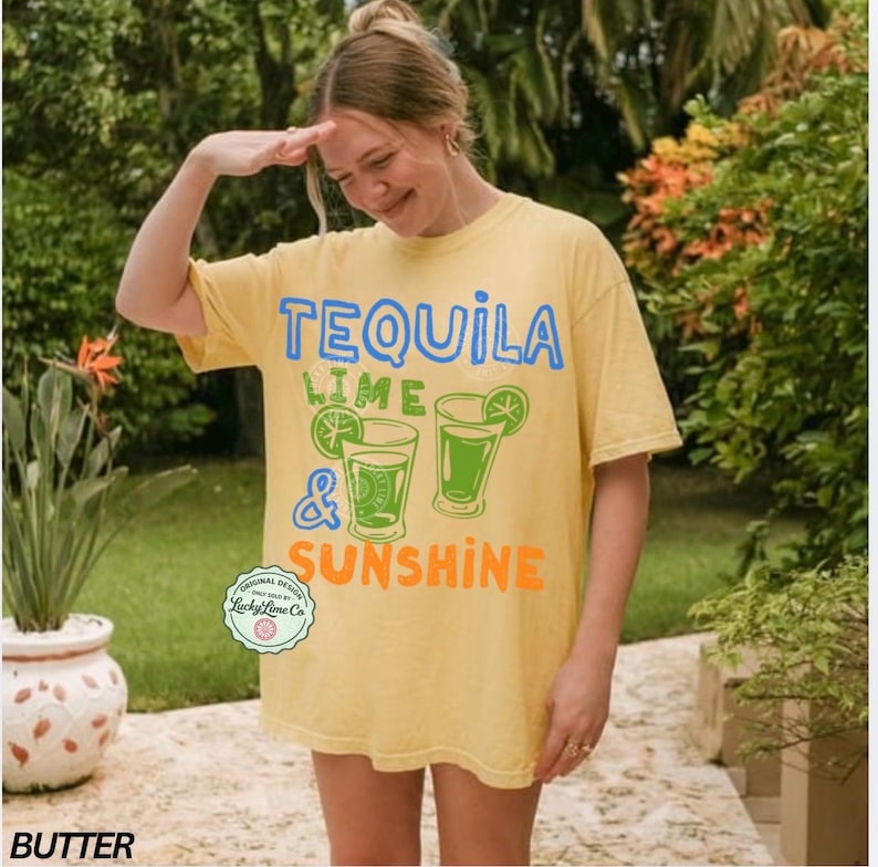 May include: A butter yellow t-shirt with the words "TEQUILA LIME & SUNSHINE" in blue, green, and orange. The shirt features two cocktail glasses with lime slices. The t-shirt is a casual, oversized fit.