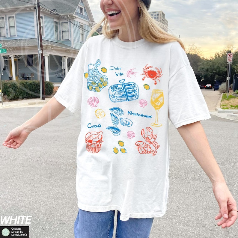 May include: White t-shirt with a colorful graphic design featuring various seafood items, including a net bag of lemons, a can of sardines, oysters, a glass of wine, and a lobster. The text "Dolce Vita" and "Mediterranean" are included in the design.