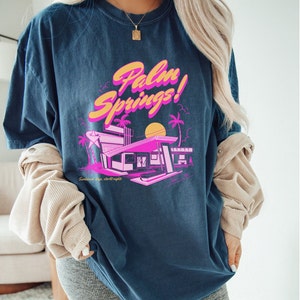 May include: A navy blue t-shirt featuring a graphic print of a pink and yellow building with palm trees and a sunset. The text "Palm Springs!" is printed in pink and yellow on the shirt. The text "Sunless days, starlit nights" is printed in white below the graphic.