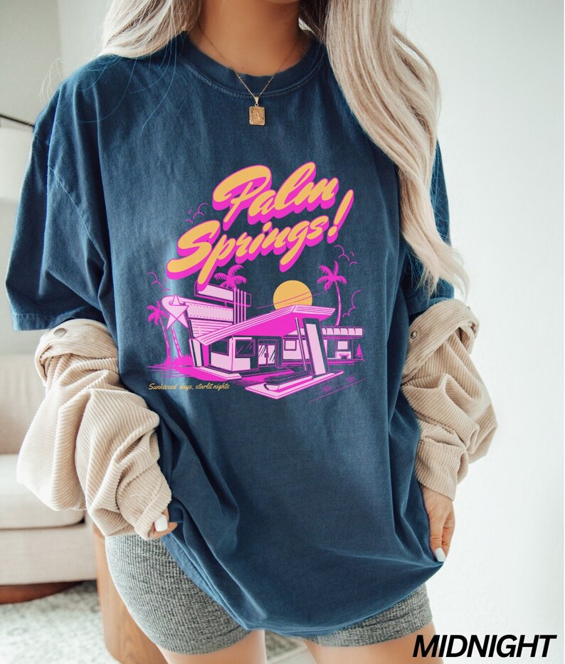 May include: A navy blue t-shirt featuring a graphic print of a pink and yellow building with palm trees and a sunset. The text "Palm Springs!" is printed in pink and yellow on the shirt. The text "Sunless days, starlit nights" is printed in white below the graphic.