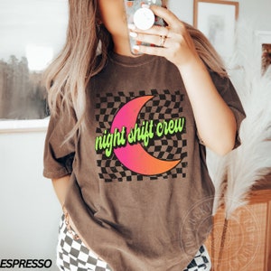 May include: A brown t-shirt with a graphic of a crescent moon on a checkered background. The text "night shift crew" is written in green on the moon.
