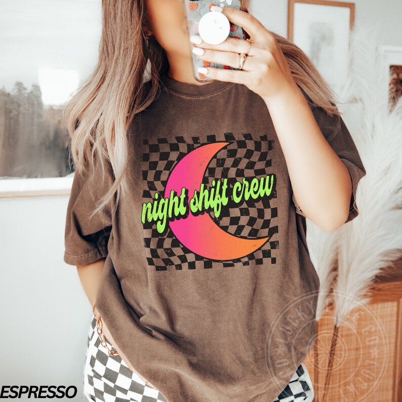 May include: A brown t-shirt with a graphic of a crescent moon on a checkered background. The text "night shift crew" is written in green on the moon.