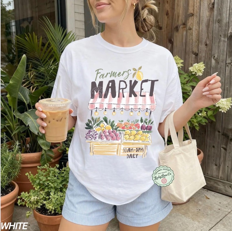 May include: White t-shirt with a colorful Farmers Market graphic. The design features a market stall with fruits and vegetables, and the text "Farmers Market" in a stylized font. The shirt also has the text "10AM-5PM DAILY".