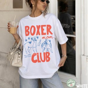 May include: A white short-sleeved t-shirt featuring the red text BOXER mom CLUB and blue line art illustrations of two boxer dogs. The design includes small paw prints and the script text Life is better with boxers. The shirt has a crew neckline.