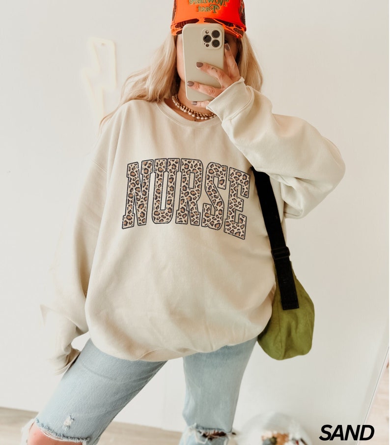 Puede incluir: Una sudadera beige con la palabra "NURSE" impresa en un dise&ntilde;o de estampado de leopardo. La sudadera la lleva puesta una persona con una gorra de b&eacute;isbol roja.