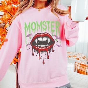 May include: A light pink sweatshirt with a graphic print of a dripping vampire mouth with fangs and the word "MOMSTER" in green letters.