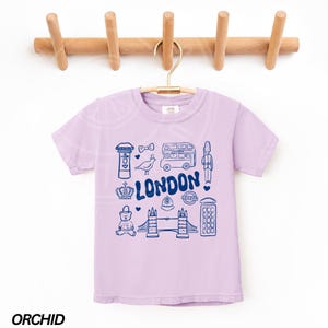 May include: A lavender coloured t-shirt with a graphic print of London landmarks in blue. The landmarks include a double-decker bus, a red telephone box, Big Ben, and Tower Bridge. The text "LONDON" is printed in blue.