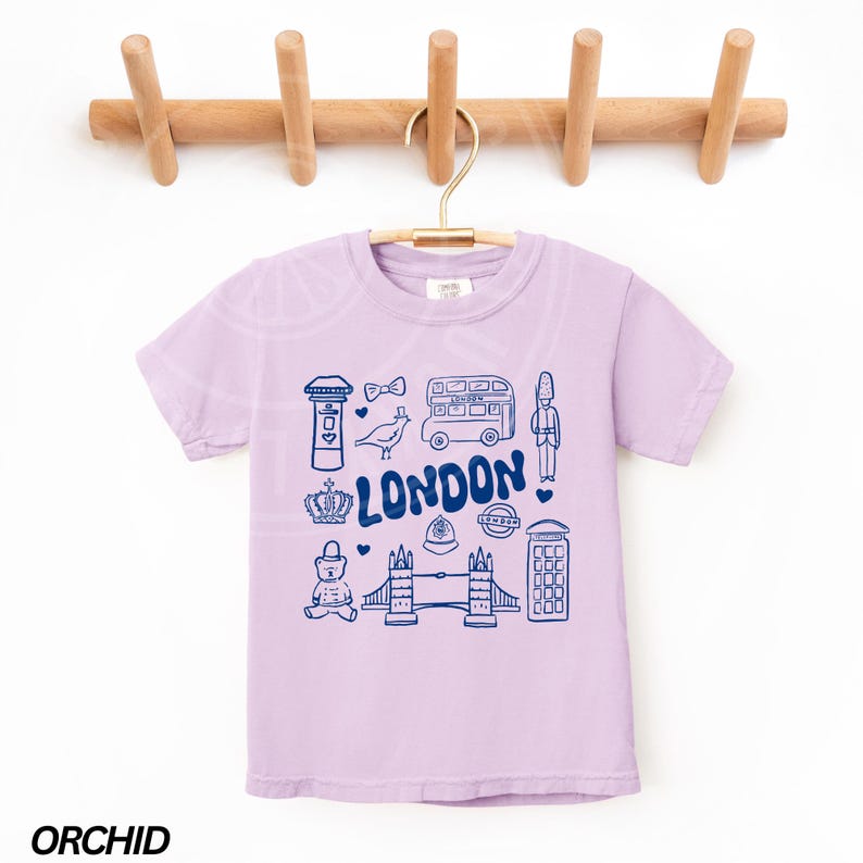 May include: A lavender coloured t-shirt with a graphic print of London landmarks in blue. The landmarks include a double-decker bus, a red telephone box, Big Ben, and Tower Bridge. The text "LONDON" is printed in blue.