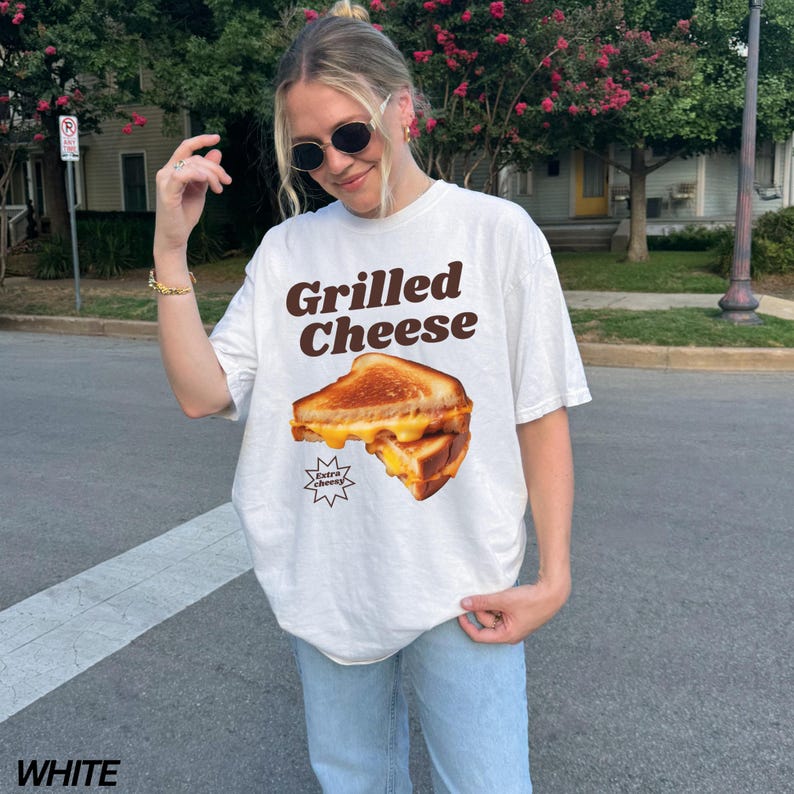 May include: White t-shirt with a graphic of a grilled cheese sandwich and the text "Grilled Cheese" in brown letters. The text "Extra Cheesy" is in a smaller font below the sandwich.