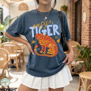 May include: Navy blue t-shirt with the text "easy TIGER" above a stylized orange and red tiger illustration. The design includes yellow star accents. The shirt is a relaxed fit.