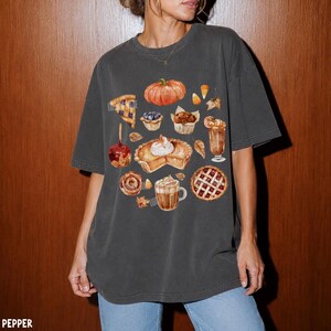 May include: A charcoal gray short-sleeve t-shirt featuring a graphic print of various autumn-themed treats, including a pumpkin, a slice of pie, a muffin, a caramel apple, a cinnamon roll, a latte, and a lattice-crust pie with scattered candy corn.