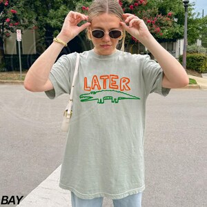 May include: A light green t-shirt with a cartoon alligator and the word "LATER" printed in orange and green.