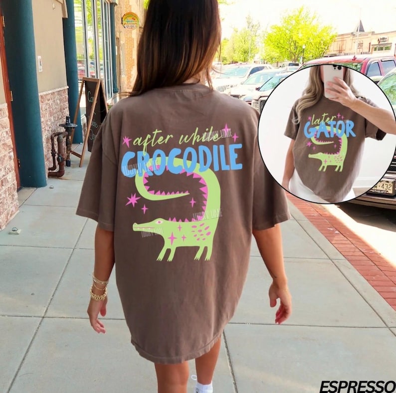 May include: Espresso-colored t-shirt with a graphic of a green alligator and the text "after while CROCODILE". The design includes pink stars and a light blue "CROCODILE" text. The shirt is a casual, oversized fit.