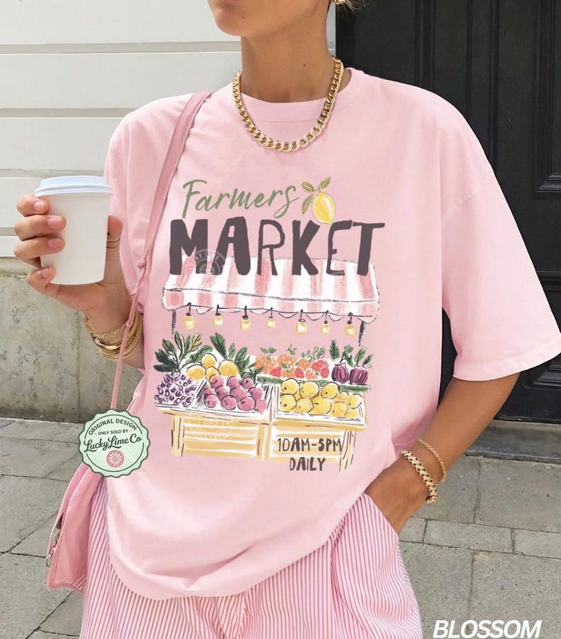 May include: A light pink t-shirt with a "Farmers Market" graphic featuring a fruit stand with various fruits and vegetables. The shirt also has the text "10AM-5PM DAILY". The shirt is part of the "Blossom" collection.