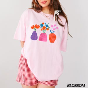 May include: A pink t-shirt with a graphic of three colorful vases with flowers. The vases are in a row and have different shapes and patterns. The flowers are in various colors, including red, blue, and pink.
