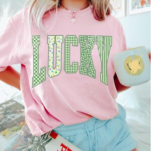 May include: A light pink sweatshirt with the word "LUCKY" in green, patterned lettering. The patterns include checkered, polka dot, and striped designs. The person is wearing light blue shorts and holding a mug.
