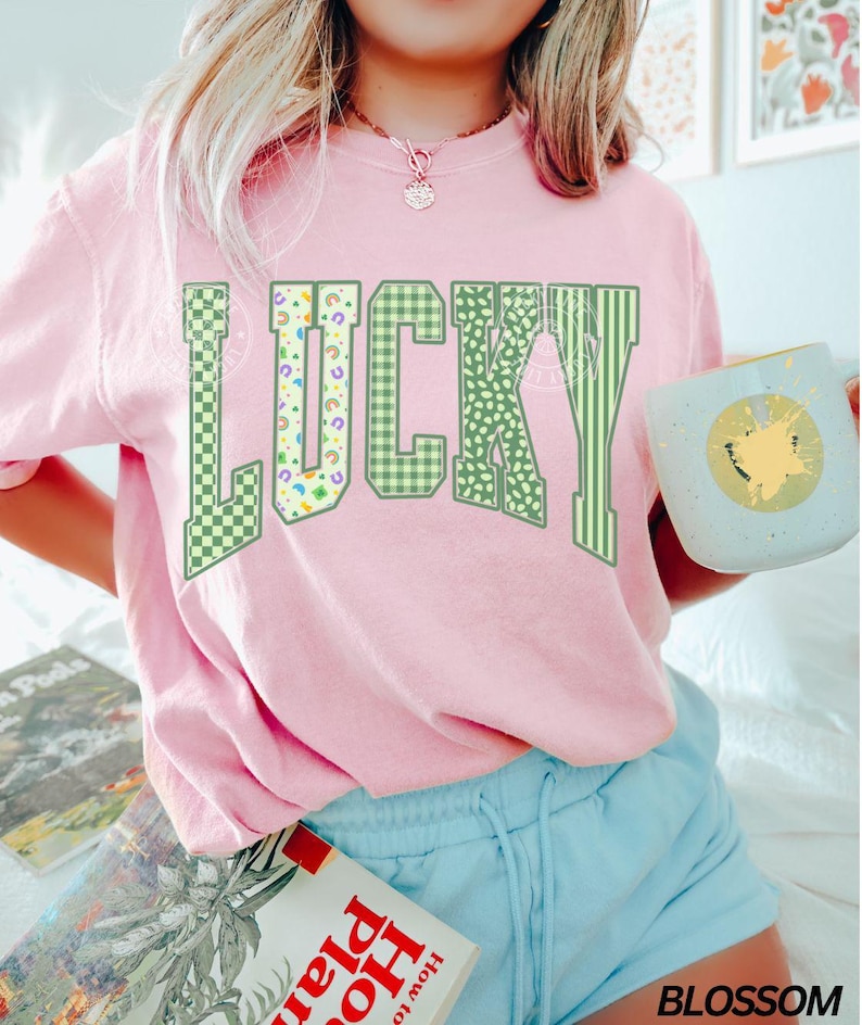 May include: A light pink sweatshirt with the word "LUCKY" in green, patterned lettering. The patterns include checkered, polka dot, and striped designs. The person is wearing light blue shorts and holding a mug.