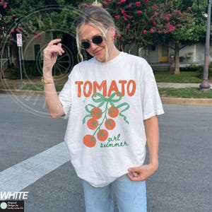 May include: White t-shirt with the word "TOMATO" in orange, a green bow, and orange tomatoes. The text "girl summer" is written in green below the tomatoes. The shirt is part of the "Original Design by Lucky Lime Co" collection.