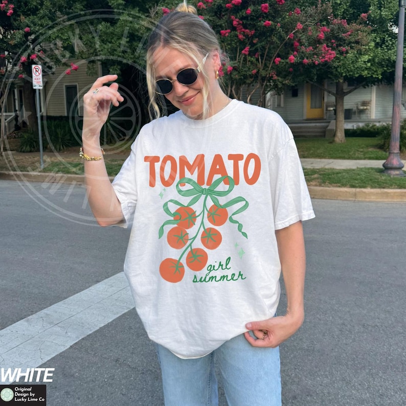 May include: White t-shirt with the word "TOMATO" in orange, a green bow, and orange tomatoes. The text "girl summer" is written in green below the tomatoes. The shirt is part of the "Original Design by Lucky Lime Co" collection.