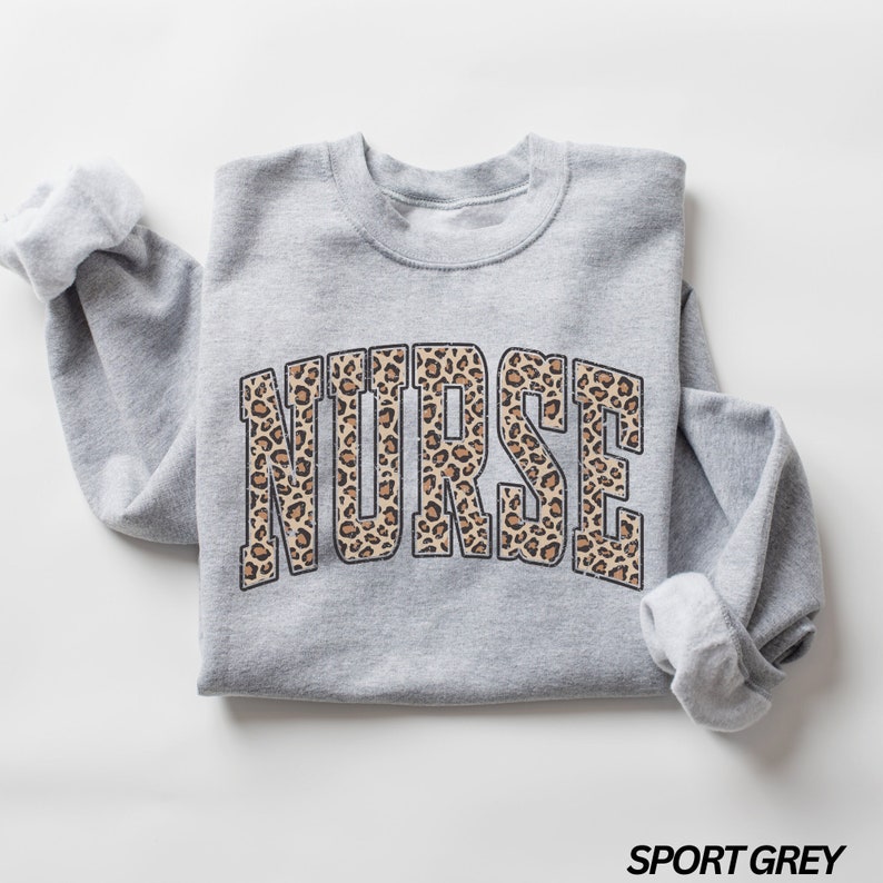 Puede incluir: Una sudadera gris con la palabra "NURSE" impresa en un estampado de leopardo. La sudadera est&aacute; doblada y colocada sobre un fondo blanco. El texto "SPORT GREY" est&aacute; impreso en negro en la parte inferior de la imagen.