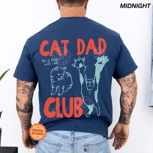 Peut inclure: T-shirt bleu nuit avec l'inscription "CAT DAD CLUB" en orange. Le t-shirt pr&eacute;sente des illustrations de chats et la phrase "life is better with cats". Un logo circulaire "Lucky Line" est &eacute;galement visible.