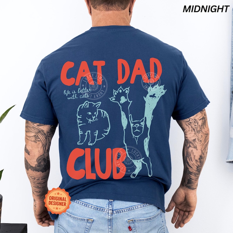 Peut inclure: T-shirt bleu nuit avec l'inscription "CAT DAD CLUB" en orange. Le t-shirt pr&eacute;sente des illustrations de chats et la phrase "life is better with cats". Un logo circulaire "Lucky Line" est &eacute;galement visible.