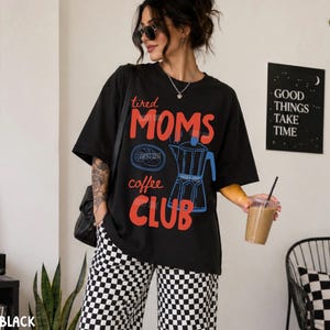 May include: A black short-sleeve t-shirt featuring the red text "tired MOMS" and "coffee CLUB" with a blue graphic of a moka pot and a small plate. The shirt is paired with black and white checkered pants, creating a casual and comfortable outfit.