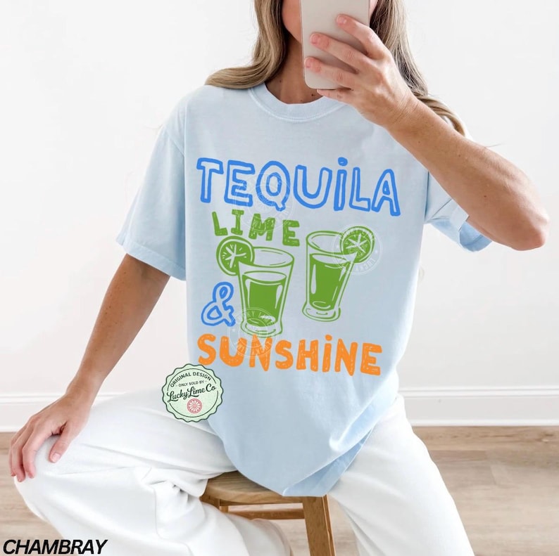May include: Pale blue t-shirt with the text "TEQUILA LIME & SUNSHINE" in blue, green, and orange. The design includes two cocktail glasses with lime slices. A casual, short-sleeved top.