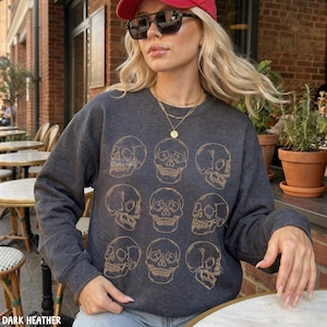 May include: A dark charcoal gray crewneck sweatshirt featuring a repeating pattern of nine gold-colored line art skulls arranged in a grid. The long-sleeved garment has a ribbed neckline, cuffs, and hem, and is worn with a gold layered necklace.