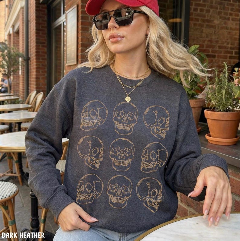 May include: A dark charcoal gray crewneck sweatshirt featuring a repeating pattern of nine gold-colored line art skulls arranged in a grid. The long-sleeved garment has a ribbed neckline, cuffs, and hem, and is worn with a gold layered necklace.