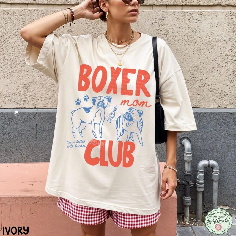 May include: An ivory oversized t-shirt featuring the red text BOXER, the blue script text mom, and the red text CLUB. The graphic includes two blue line-art boxer dogs, blue paw prints, and the text Life is better with boxers in blue script.