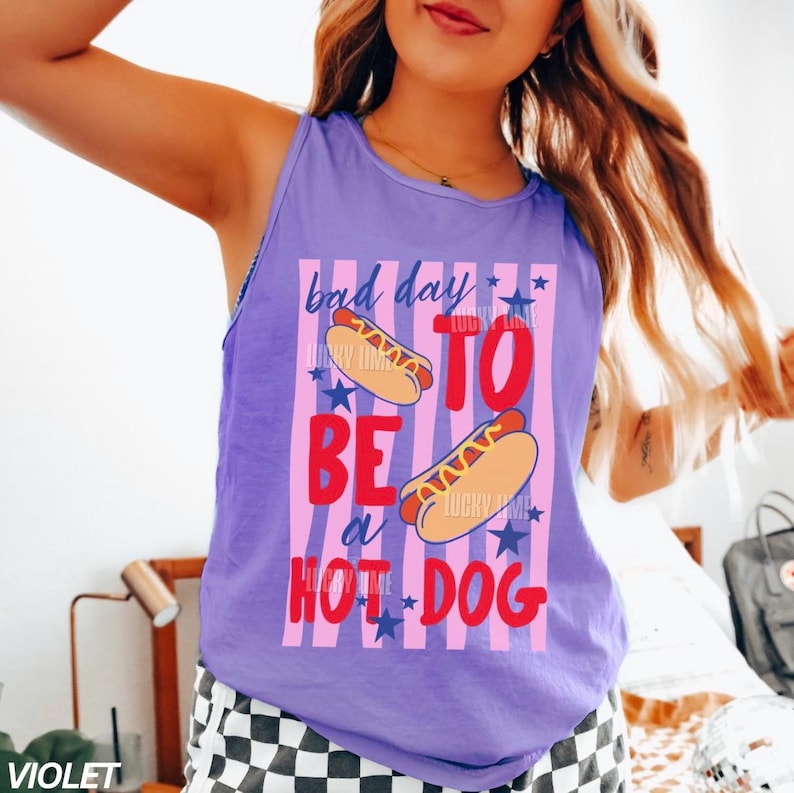 Op de afbeelding: Een violette tanktop met de tekst "bad day to be a hot dog" in rood en wit. Het ontwerp bevat illustraties van hotdogs en blauwe sterren. Een casual zomerkledingstuk.