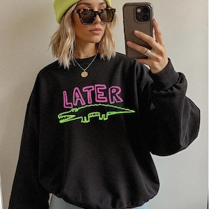 May include: Black sweatshirt featuring the word "LATER" in pink and a green alligator design. The sweatshirt is worn with a yellow beanie and sunglasses. The word "BLACK" is printed in white on the bottom right.