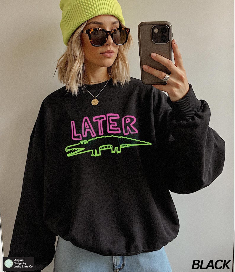 May include: Black sweatshirt featuring the word "LATER" in pink and a green alligator design. The sweatshirt is worn with a yellow beanie and sunglasses. The word "BLACK" is printed in white on the bottom right.