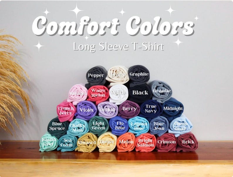 Może przedstawiać: Kolekcja zwiniętych koszulek z długim rękawem w r&oacute;żnych kolorach, ułożonych w kształt piramidy. Na g&oacute;rze widnieje napis Comfort Colors Long Sleeve T-Shirt. Każda rolka jest oznaczona nazwami kolor&oacute;w, takimi jak Pepper, Ivory, Graphite, Blossom, Watermelon, White, Black, Granite, Crunchberry, Violet, Orchid, Berry, True Navy, Midnight, Blue Spruce, Chalky Mint, Light Green, Flo Blue, Lagoon Blue, Blue Jean, Chambray, Island Reef, Seafoam, Mustard, Butter, Melon, Bright Salmon, Crimson i Brick.