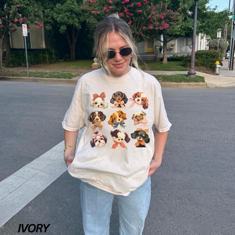 May include: A woman wearing a cream-colored t-shirt with a graphic of nine different dogs wearing bows. The t-shirt has a relaxed fit and is made of a soft, comfortable fabric. The word "IVORY" is printed in the bottom left corner of the image.
