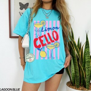 May include: Lagoon blue t-shirt with a graphic design featuring the words "Limon Cello" and illustrations of cocktails and lemons. The design is set against pink and blue vertical stripes. The shirt has a relaxed fit.