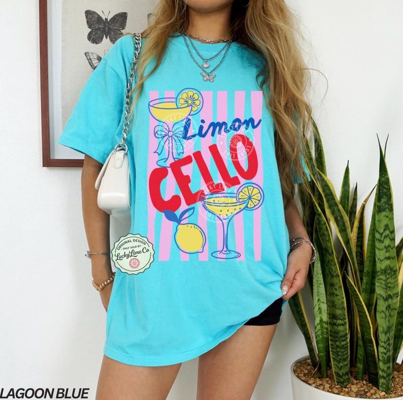 May include: Lagoon blue t-shirt with a graphic design featuring the words "Limon Cello" and illustrations of cocktails and lemons. The design is set against pink and blue vertical stripes. The shirt has a relaxed fit.