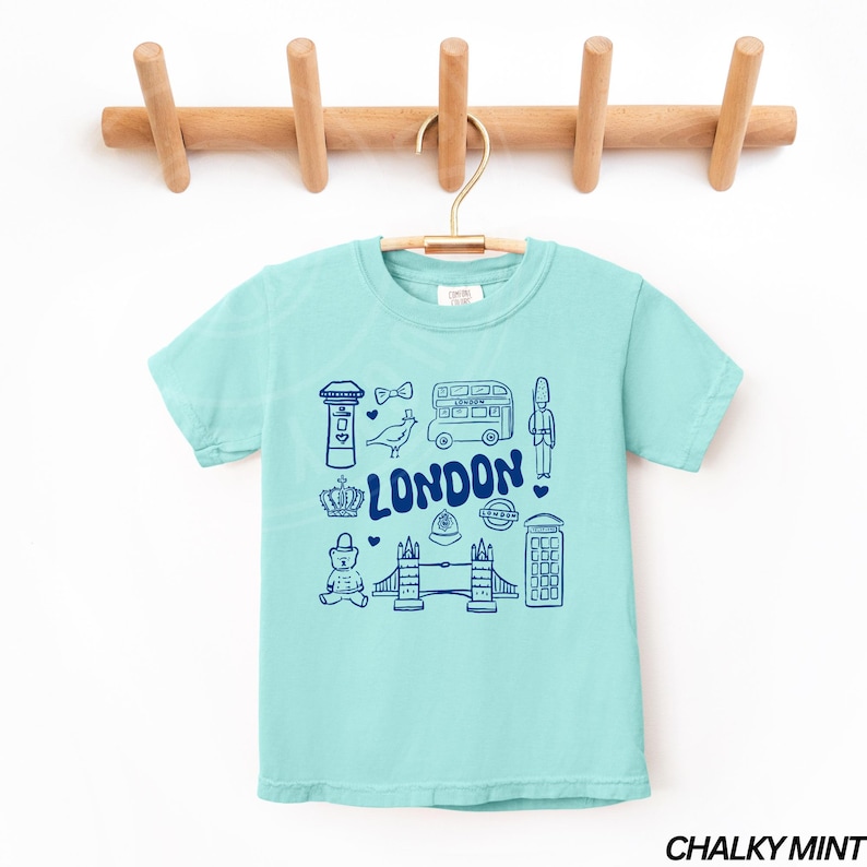 May include: A light blue t-shirt featuring a graphic print of London landmarks in blue. The landmarks include a double-decker bus, a red telephone box, Big Ben, and Tower Bridge. The text "LONDON" is printed in blue on the shirt.